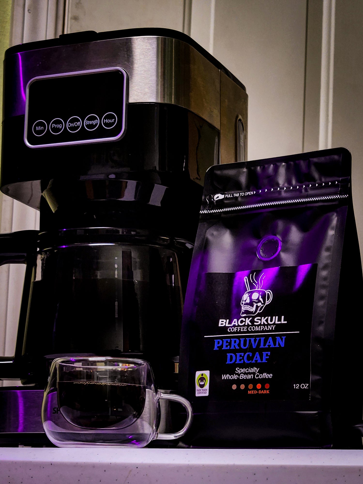 Black Skull Coffee Company Peruvian Decaf package sitting on a desk with a clear glass coffee cup and a full coffee cup