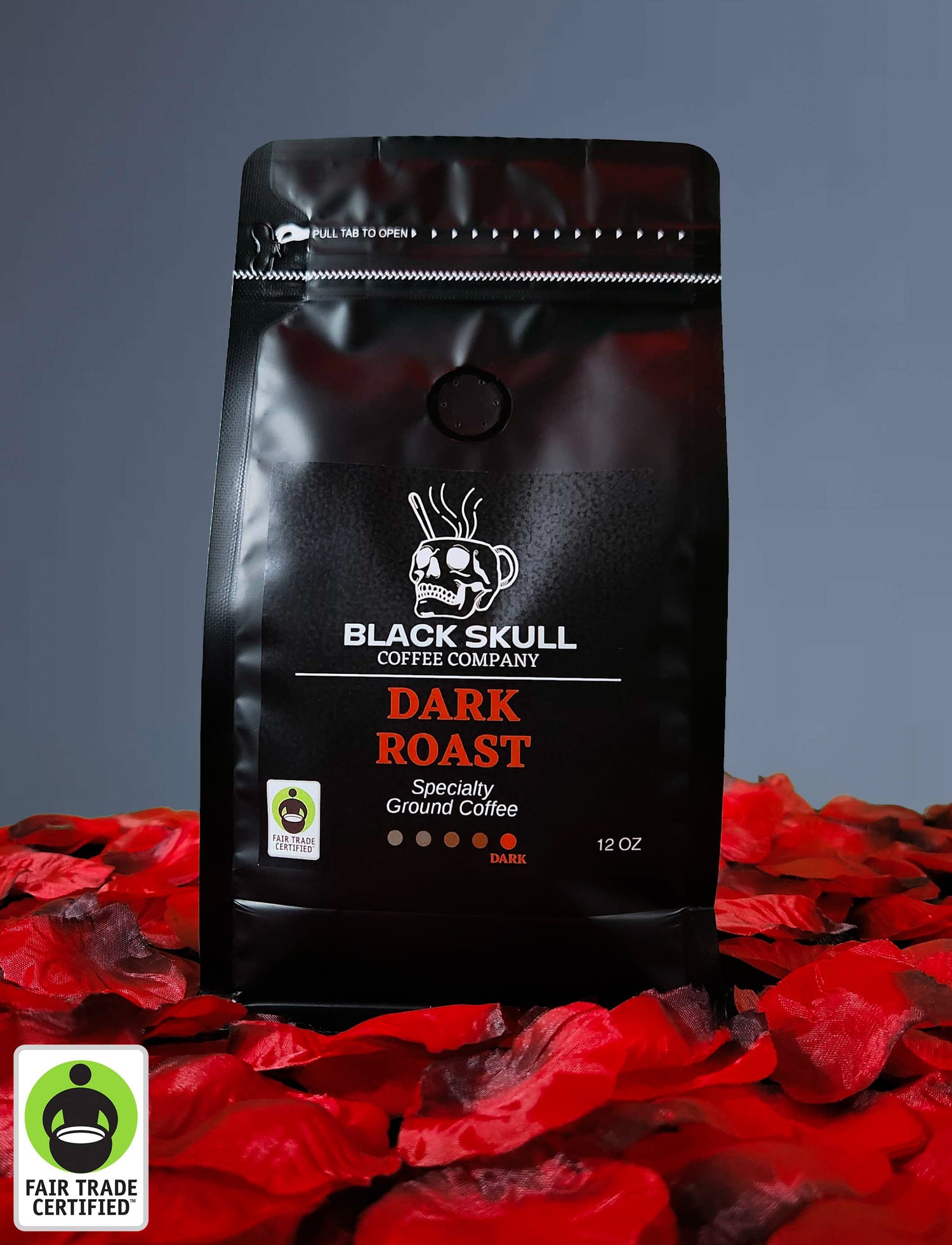 Black Skull Coffee Company Specialty Dark Roast Coffee 12oz ground coffee package on a gray background with red rose petals and Fair Trade USA Logo.